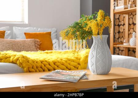 Vase mit Mimosenblumen und Zeitschriften auf dem Tisch im Schlafzimmer, Nahaufnahme Stockfoto