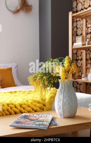 Vase mit Mimosenblumen und Zeitschriften auf dem Tisch im Schlafzimmer, Nahaufnahme Stockfoto