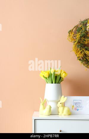 Blumenstrauß, Dekor und Grußkarte mit Text HAPPY EASTER auf Kommode in der Nähe der farbigen Wand Stockfoto