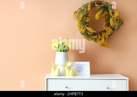 Blumenstrauß, Dekor und Grußkarte mit Text HAPPY EASTER auf Kommode in der Nähe der farbigen Wand Stockfoto