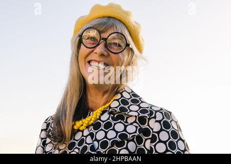 Fröhliche ältere Frau, die fröhlich lächelt, während sie im Freien steht. Glückselige ältere Frau in farbenfroher Freizeitkleidung im Sommer. Sorglos reif Stockfoto