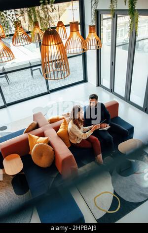 Blick von oben auf zwei Geschäftsleute, die in einer Bürolobby arbeiten. Zwei moderne Geschäftsleute, die ein digitales Tablet nutzen, während sie eine Diskussion führen. Zwei junge Stockfoto