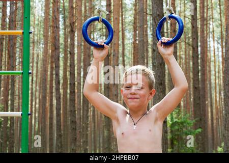 Porträt eines niedlichen Jungen, der sich vor dem Hintergrund eines Waldes auf Turnringen in der Natur hochzieht, Outdoor-Kurse Stockfoto