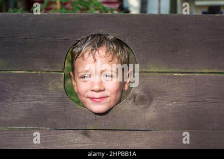 Ein süßer kaukasischer Junge schaut durch ein Loch in einem Holzzaun und lächelt. Stockfoto