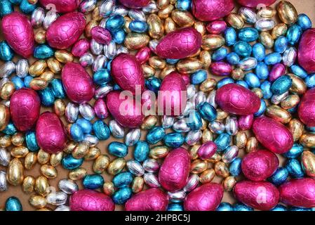 Kleine rosafarbene, blaue, silberne und goldene Schokoladen-ostereier, mit großen rosafarbenen Eiern auf hellem Holzgrund. Stockfoto