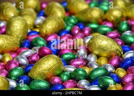 Kleine rosafarbene, blaue, grüne und silberne Schokoladen-ostereier mit größeren gelben Eiern auf hellem Holzgrund. Stockfoto