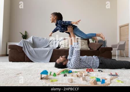 Glücklicher junger afroamerikanischer Vater, der in der Luft freudige Tochter hebt. Stockfoto