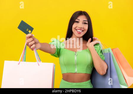 Einfaches Einkaufen. Fröhliche Dame Zeigt Kreditkarte Nahaufnahme Der Kamera Hält Helle Einkaufstaschen, Glückliche Frau Trägt Einkäufe, Erhielt Cashback Bonus, Pos Stockfoto