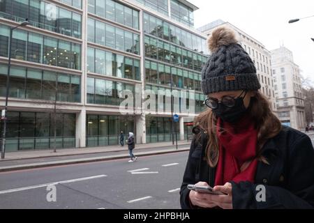 Frau trägt Wollmütze, Brille und Gesichtsmaske mit Handy auf einer Londoner Straße Stockfoto