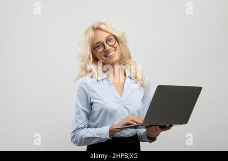 Porträt einer aufgeregten jungen Geschäftsfrau, die einen Laptop in der Hand hält, am Computer arbeitet und die Kamera anlächelt Stockfoto