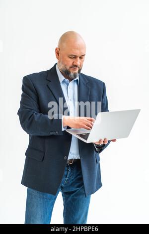 Vertikales Foto eines reifen Mannes, der auf weißem Hintergrund bleibt. Arbeiten mit einem Laptop. Tragen Sie blaue Jacke, Hemd, Jeans. Nahaufnahme Stockfoto