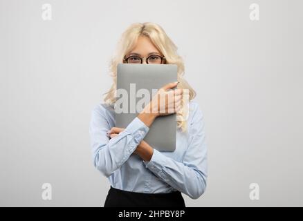 Erstaunte junge Geschäftsfrau, die das Gesicht mit einem Laptop bedeckt, formelle Kleidung trägt und auf hellem Studiohintergrund posiert Stockfoto