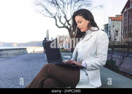 Junge kaukasische Frau, die mit einem Laptop an einer Strandpromenade arbeitet. Stockfoto