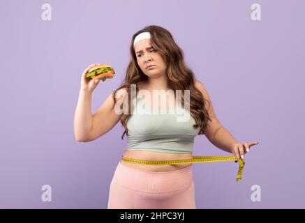 Traurige europäische Millennial plus size Dame in Sportbekleidung misst Taille mit Maßband und schaut Burger, isoliert auf violettem Hintergrund, Studio. Fa Stockfoto