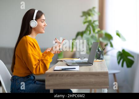 Glückliche junge kaukasische Frau, die Kopfhörer trägt, Laptop benutzt, von zu Hause aus arbeitet oder online studiert, leerer Raum Stockfoto