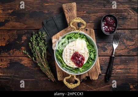 Gebackener Camembert Brie-Käse mit Cranberry-Sauce und garniert mit Rucola-Salat in einer Pfanne. Holzhintergrund. Draufsicht Stockfoto