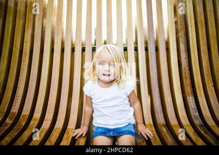 Nettes blondes Mädchen, das auf einer Holzbank im Park sitzt. Stockfoto