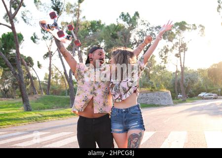 Junge, coole tätowierte Paare brüllen, erheben ihre Arme und lachen mit einem Skateboard. Stockfoto