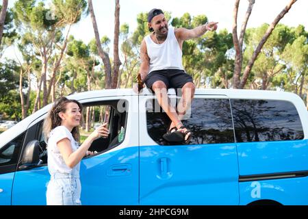 Das junge tätowierte Paar sitzt auf dem Dach des Wagens, trinkt Bier, zeigt und lacht. Stockfoto