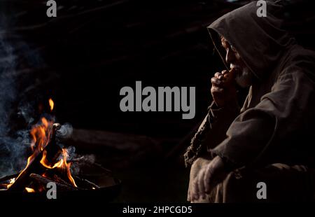 Alter armer Mann, der im Mittelalter mit einer Flöte am Feuer saß Stockfoto