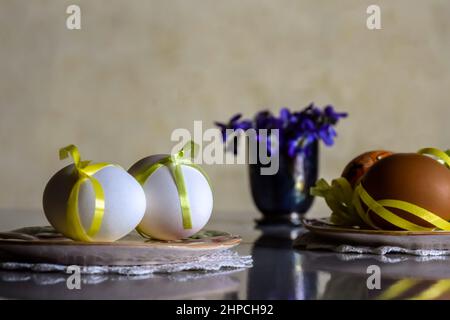 Ostern: Zwei Keramikplatten mit weißen und braunen Eiern, verziert mit Bändern, und ein Strauß blauer Waldblumen in einer kleinen Vase spiegeln sich Stockfoto