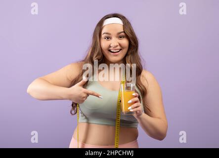 Fröhliche lächelnde und große europäische Millennials-Dame in Sportswear mit Messband zeigt den Finger auf ein Glas Orangensaft Stockfoto