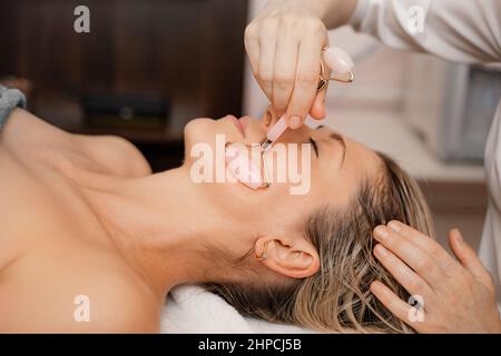 Attraktive Frau Gesichtsmassage Gua Sha rosa Lifting Stone Roller. Nahaufnahme. Schönheitssalon. Anti-Aging-Therapie. Pflege Stockfoto