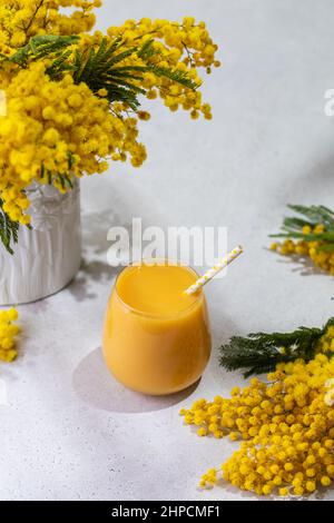 Orangensaft, Zitrus-Smoothie-Glas auf Frühlingshintergrund, mit Mimosa-Blumenvase, hartem, umweltfreundlichem Cocktailstroh Stockfoto