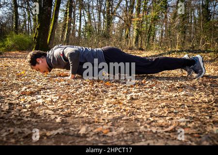 Der Läufer macht Liegestütze im Wald. Der junge Mann mit den blauen Augen und dem Schnurrbart macht Liegestütze auf dem Boden, während er der Musik zuhört Stockfoto