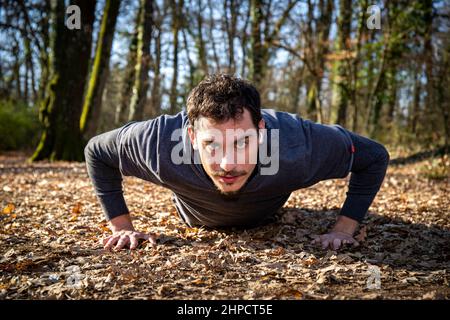 Runner macht Liegestütze im Wald. Der junge Mann mit den blauen Augen und dem Schnurrbart macht Liegestütze auf dem Boden, während er der Musik zuhört. Stockfoto