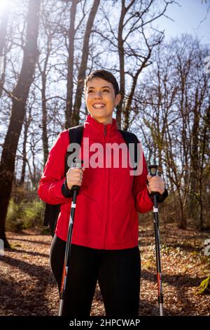 Die lächelnde junge Frau wandert im Wald. Die Frau hat kurze Haare, trägt eine rote Jacke und verwendet Trekkingstöcke. Reise- und Outdoor-Konzept. Stockfoto