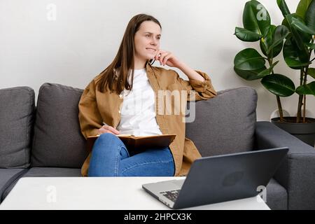 Glückliche junge Frau sitzen auf dem Sofa Studie online am Computer Notizen schreiben Träumen oder Denken. Lächelnde kaukasische Frauen arbeiten zu Hause fern auf dem Laptop Stockfoto