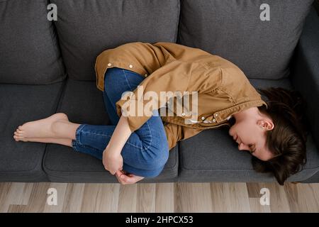 Draufsicht auf müde unglückliche junge Frau, die auf der Couch zusammengerollt liegt, leidet an Menstruationskrämpfen. Ungeplante Schwangerschaft und Abtreibung, versuchen zu schlafen Stockfoto