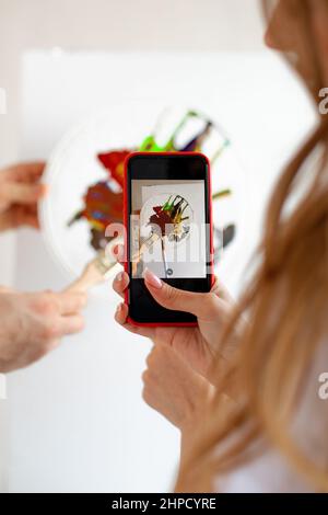 Nicht erkennbare Frau mit langen Haaren, die mit dem Handy Aquarellpalette mit Bürsten auf unbeschriftete Leinwand fotografiert. Stockfoto