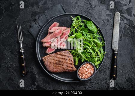 BBQ gegrilltes Rock Steak auf einem Teller mit grünem Salat. Schwarzer Hintergrund. Draufsicht Stockfoto