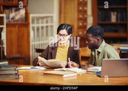 In warmen Farbtönen gehaltene Darstellung einer lächelnden erwachsenen Frau, die in einer College-Bibliothek mit einer afroamerikanischen Freundin studiert Stockfoto