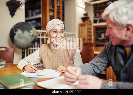 Porträt einer lächelnden muslimischen Frau mit Kopfbedeckung während des Studiums in der Universitätsbibliothek bei einem Senior Professor Stockfoto