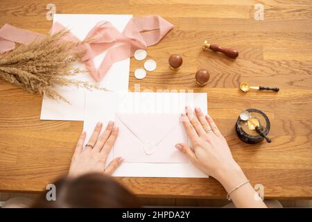 Zugeschnittenes Foto von Frau Hände, die Grußkarte Umschlag Handwerk, schmücken mit rosa Band, Stempeln. Oben. Cerealien auf dem Schreibtisch. Stockfoto