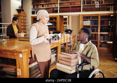 Porträt eines jungen afroamerikanischen Mannes im Rollstuhl, der Stapel Bücher hält und mit einem Freund in der Universitätsbibliothek plaudert Stockfoto