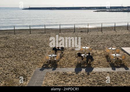 Blick auf die Küste mit Paaren, die an einer Strandbar sitzen, und dem Hafen im Hintergrund an einem sonnigen Wintertag, Sanremo, Ligurien, Italien Stockfoto