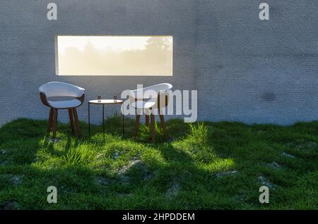 Zwei Stühle, Tisch und Wassergläser in der Nähe des Fensters mit hellem Sonnenlicht, das durch eine Betonwand im Grass and Rocks Field scheint. Stockfoto