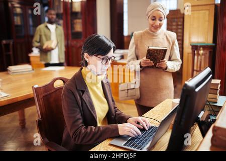Porträt einer weiblichen Bibliothekarin mit einem Laptop, während sie Studenten in der Universitätsbibliothek hilft Stockfoto