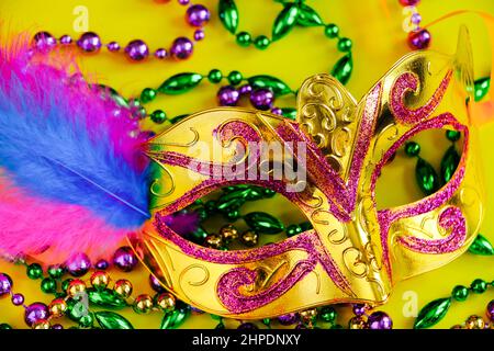 Karnevalsmaske mit Federn auf gelbem Hintergrund. Bunte Perlen in grün, Gold und lila. Symbol „Mardi Gras“ oder „Fat Tuesday“. Stockfoto