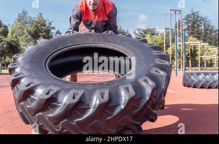 Junger Mann hebt und schiebt einen riesigen Reifen und trainiert mit funktionellem Training im Freien Stockfoto