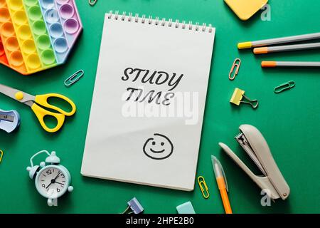 Notizbuch auf grünem Hintergrund mit Text Studienzeit.Farbig verschiedene Schulmaterialien und ein Wecker auf grünem Papierhintergrund. Stockfoto
