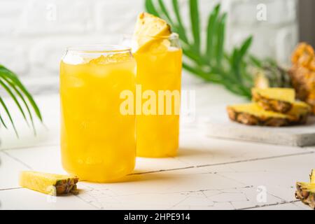 Ananascocktail oder Saft in zwei Gläsern mit Eis auf weißem Hintergrund mit Palmblättern Stockfoto