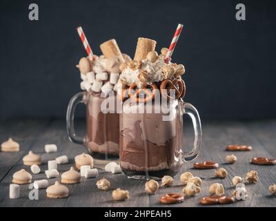 Nahaufnahme von zwei Freakshake im Einmachglas auf braunem Tisch. Ausgeflippter Milchshake mit Schokolade, Brezel, Marschmalow, Popcorn und Waffeln. Trendiges Essenskonzept - ein verrückter Shake Stockfoto