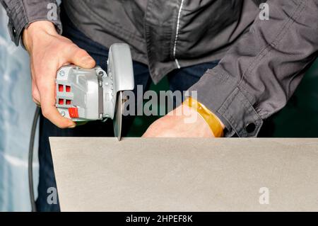 Arbeiter schneidet Fliesen. Schneiden von Keramikfliesen mit einem manuellen Elektroschleifer mit einer Diamantscheibe. Speicherplatz kopieren. Nahaufnahme. Stockfoto