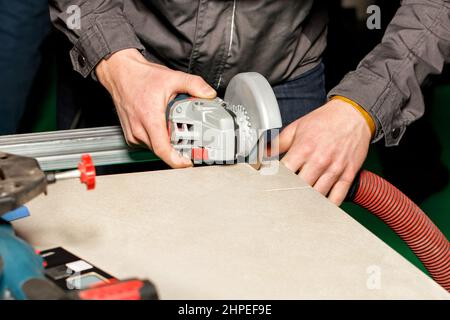 Die Hände des Meisters schnitten die Keramikplatte mit einem elektrischen Mahlwerk mit einer Diamantscheibe mit einem Staubabsaugschlauch. Speicherplatz kopieren. Nahaufnahme. Stockfoto