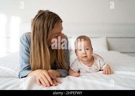 Porträt einer Mutter mit einem vier Monate alten Baby im Schlafzimmer. Junge glückliche Mutter liegt neben ihrem entzückenden kleinen Sohn, einem süßen blonden Jungen, der in einem Whit spielt Stockfoto